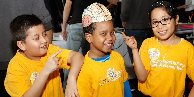 Three children in yellow "KIPP Infinity Elementary" t-shirts smiling and pointing to the brain diagram hat the middle child is wearing.