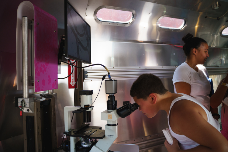 A young child peeks through a microscope on board the Columbia University Zuckerman Institute BioBus during the back-to-school event in West Harlem. Photo Credit: Henry Danner