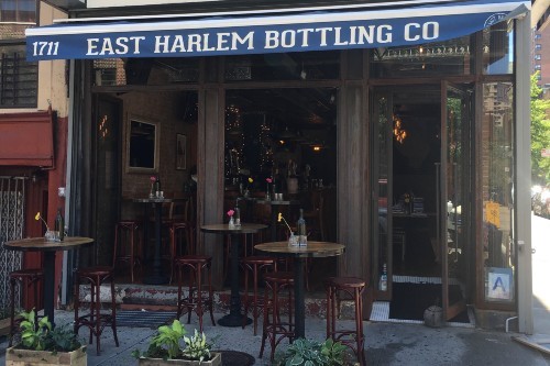 The outside of a restaurant, with bar-height tables on the sidewalk and a blue awning that says "East Harlem Bottling Co."