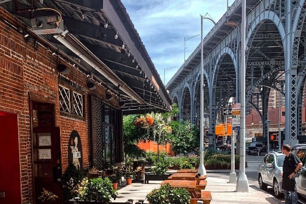 A brick building with picnic tables on the sidewalk and a raised metal viaduct to the right.