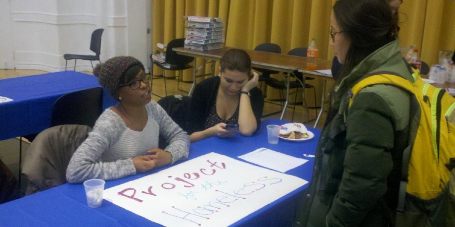 Two students speaking to a woman at an open house, with a table that has a Project for the Homeless sign between them.