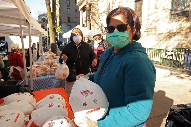 A woman in a green mask and teal sweatshirt poses with a frozen turkey in a bag.