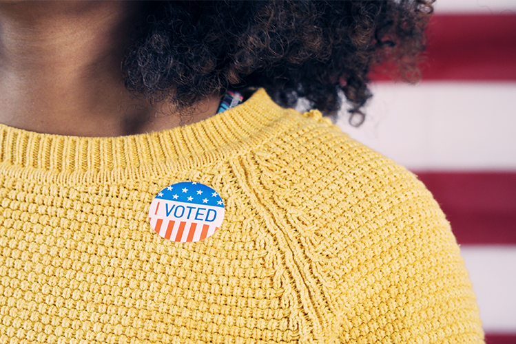 An I Voted Sticker on a person with a yellow sweater with red and white flag stripes behind.