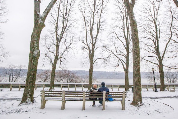 Two people sitting on a bench in a snowy park looking at the Hudson River.