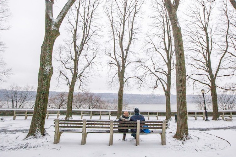 Enjoy beautiful views from Fort Tryon Park. Photo courtesy of NYC Parks. Two people sitting on a bench in a snowy park looking at the Hudson River.