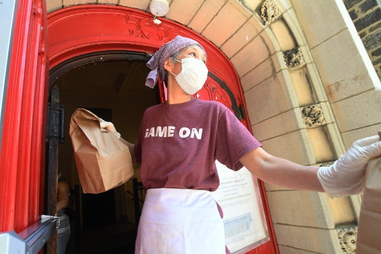 Woman wearing mask and hair cloth, in front of red door, hanging out bagged lunches.