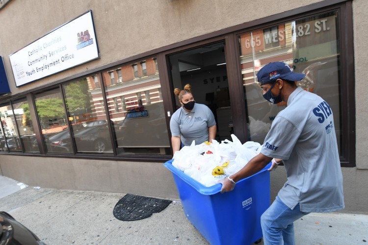 Two people rolling a large blue cart full of plastic bags of food.