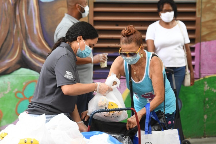 A woman wearing a surgical mask places a bag of food into a cart for another woman, who is also wearing a mask.