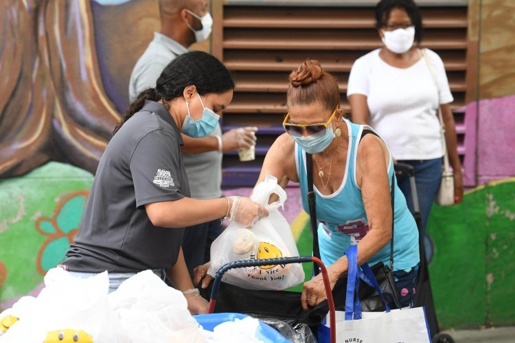 Food distribution underway at the Ecumenical Food Pantry. (Photo by Eileen Barroso.) A woman wearing a surgical mask places a bag of food into a cart for another woman, who is also wearing a mask.