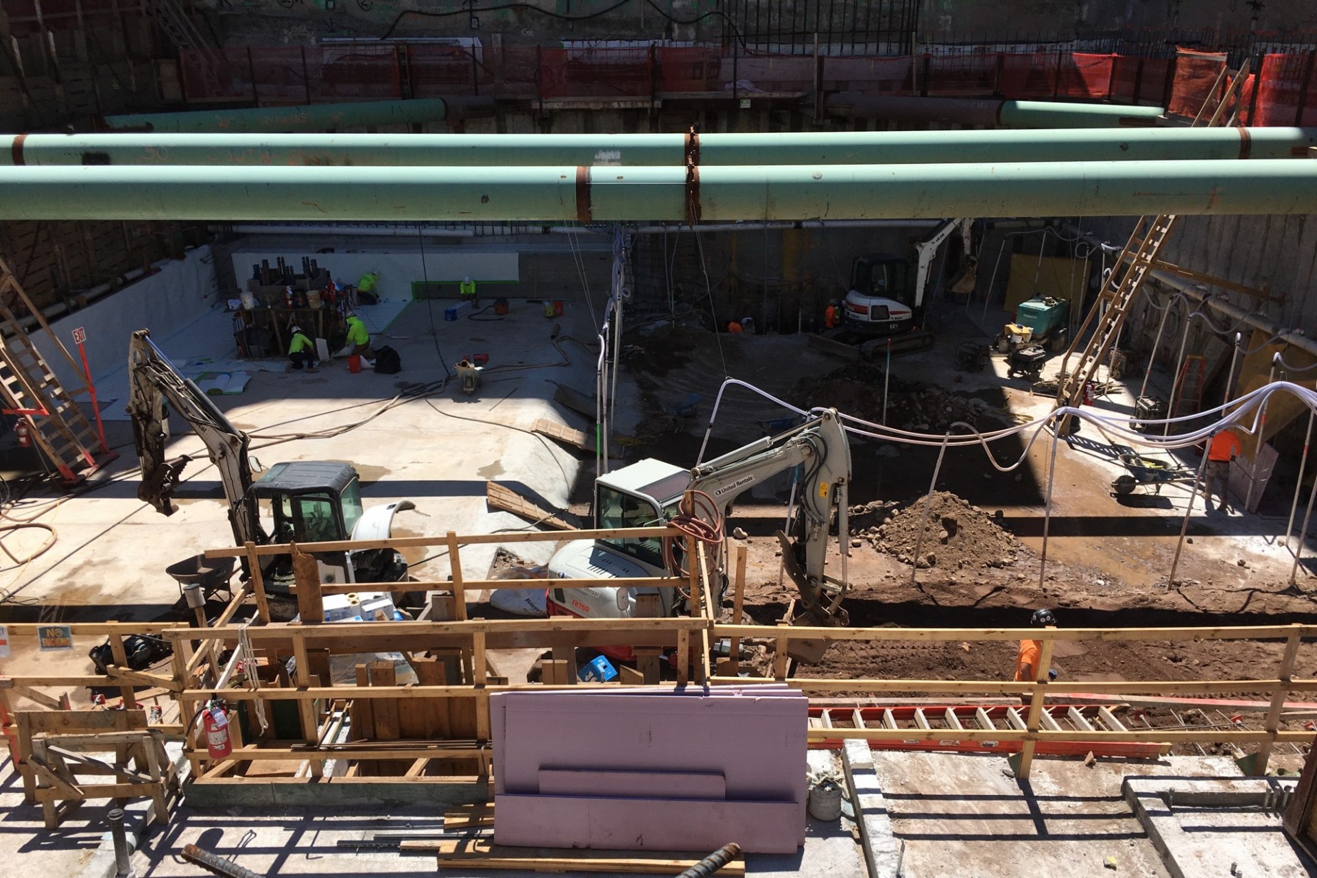 A view of the construction site from the ground level with large green pipes running overhead.