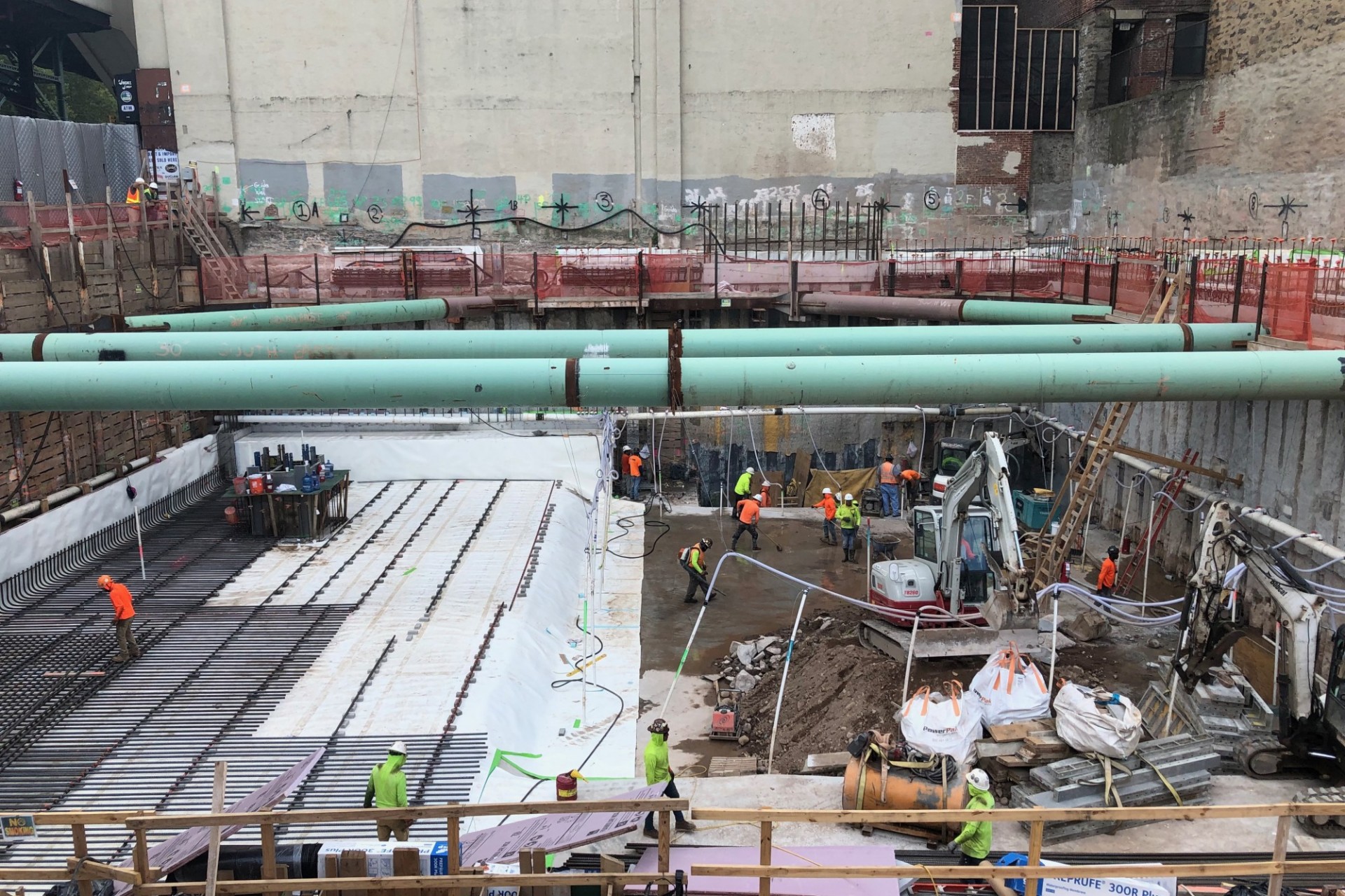 A view of the construction site from the ground level with white, thick panels of waterproofing on the left side, and large green pipes running overhead.