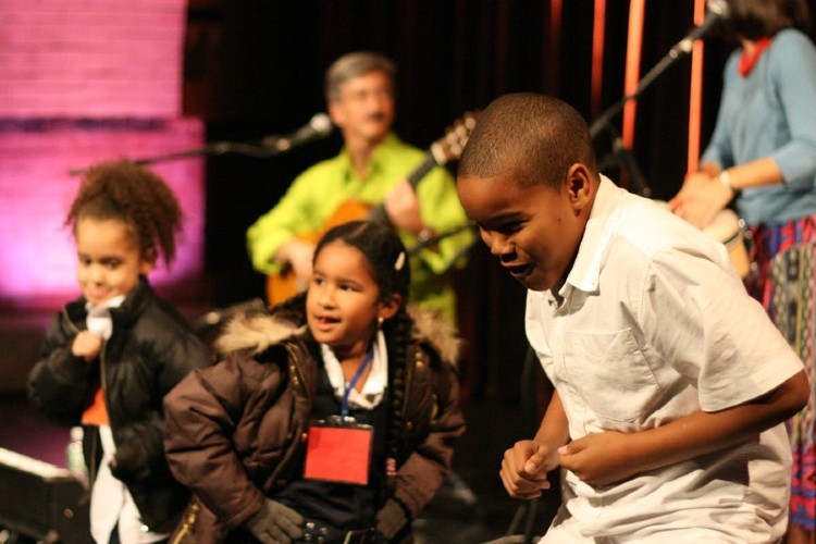 Three children performing on a stage with two adults playing instruments behind them.