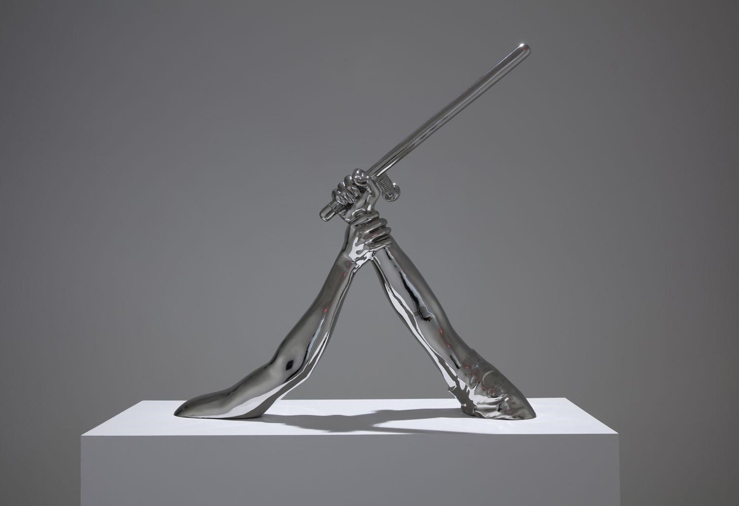 Photo of a metal sculpture by Hank Willis Thomas entitled Strike. It depicts two arms protruding from a white base. One arm is out-stretched and holding a baton-like instrument. The second arm is also reaching out, and has grasped the wrist of the other arm.