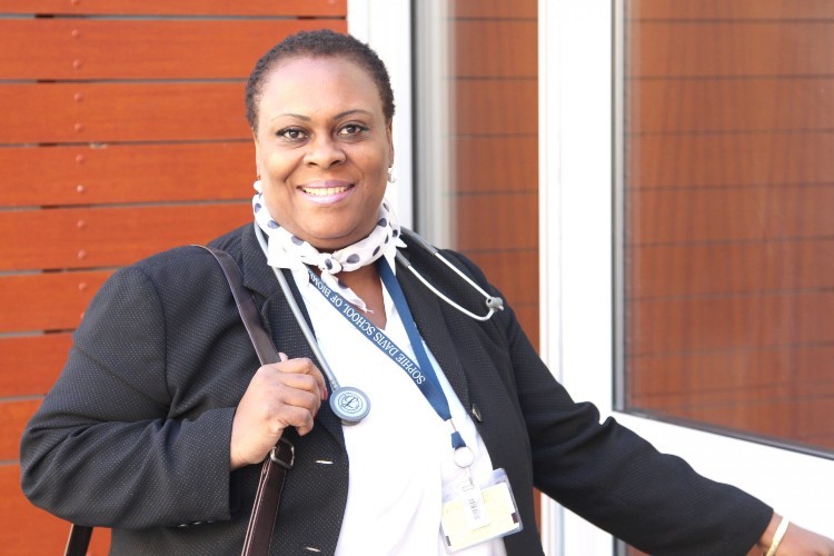 A woman with short black hair wearing a black jacket, a black and white scarf, and a stethoscope smiles at the camera.