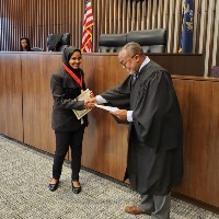 A smiling young woman holding a certificate shakes hands with a judge in a courtroom.