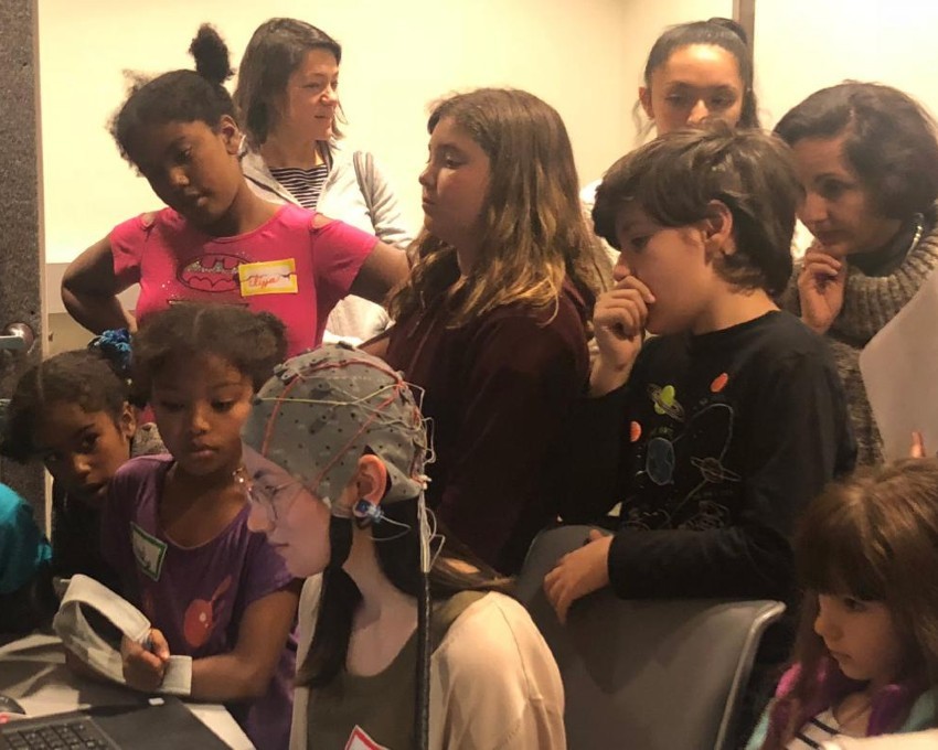 A group of children huddled around a young woman wearing a close-fitting cap covered in wires.