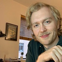 Aaron Schein: headshot of young man with blond hair and blond mustache with backdrop of window and desk.