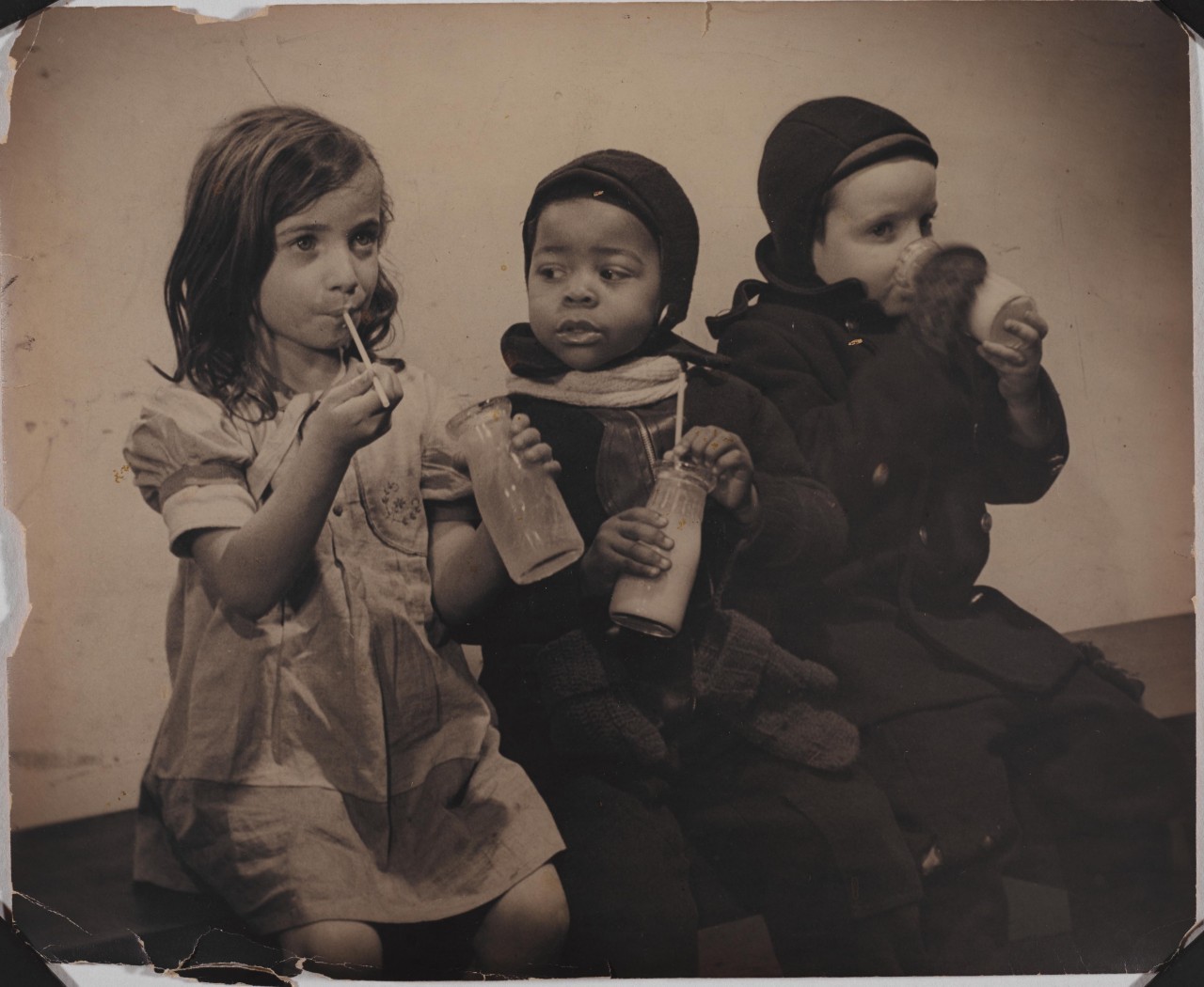 Three young children drinking milk from a glass bottle and straw.