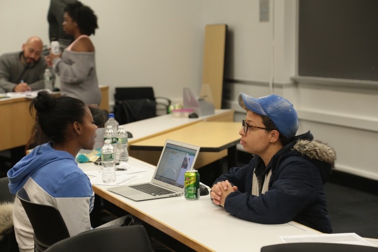 Two students in a class room discussing something while looking at a laptop