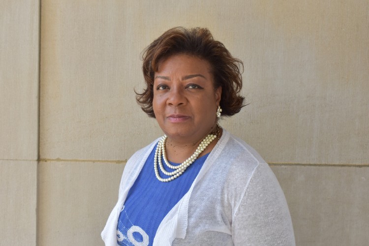 Victoria Mason-Ailey headshot: A woman standing in front of an ivory stone wall in a blue shirt and white cardigan wearing pearls.