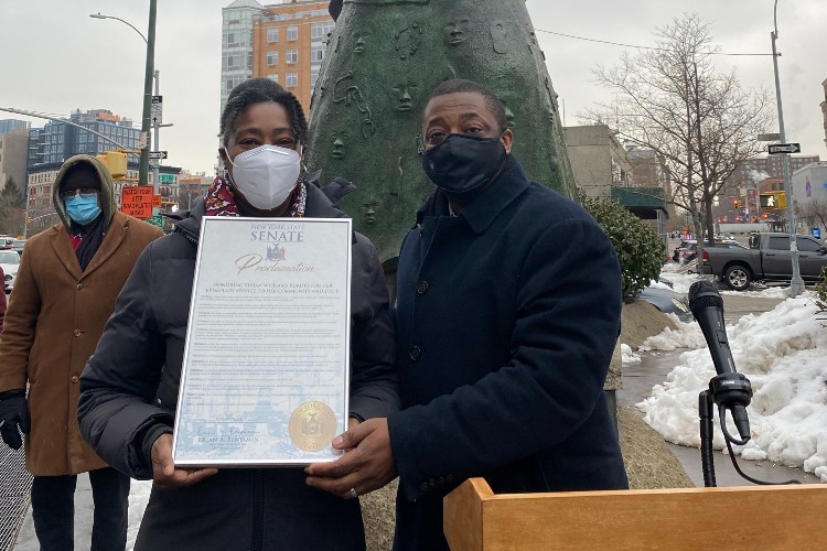 A woman and a man standing in front of a statue, both wearing masks. The woman is holding a New York State Senate proclamation.