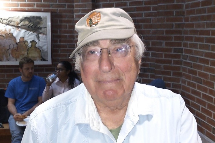 A white-haired man in a baseball cap and glasses looking at the camera.