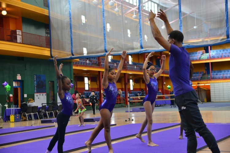 Three girls in purple leotards standing on mats with their arms raised above their heads, and a young man in front of them in a purple shirt demonstrating the pose.