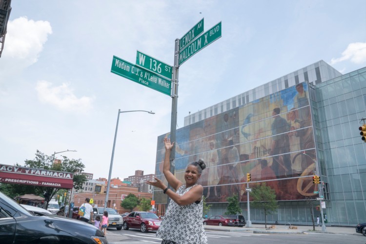 Bundles standing outside underneath the street sign marking Madam C. J. & A'Lelia Walker Place at 136th Street and Lenox Avenue.