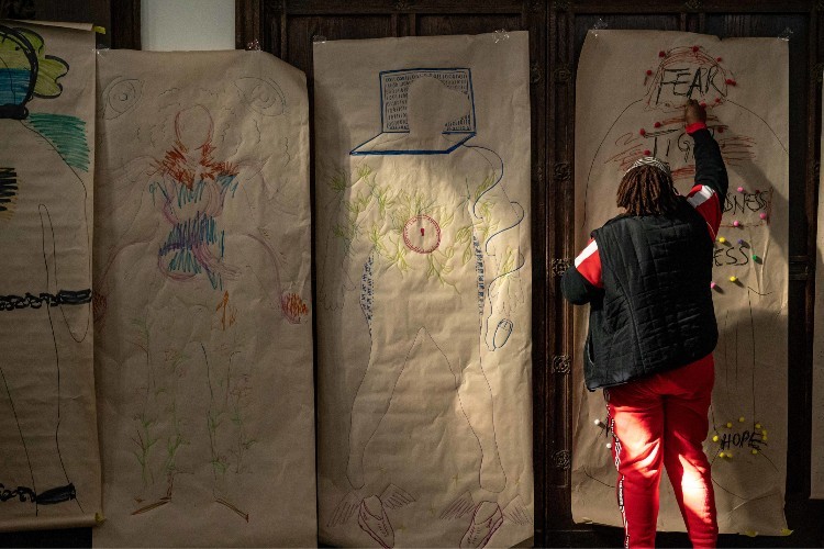 A woman stands with her back to the camera drawing on a large piece of paper that has the outline of a human on it. The paper says "fear."