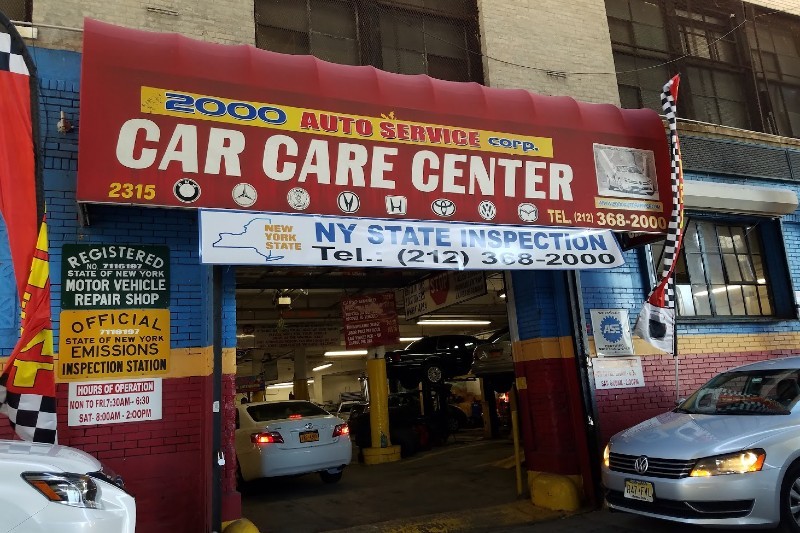 A red awning for Auto Service Car Care Center with a banner below it advertising NY State inspections and sharing their phone number. Cars are parked outside the opening to the garage.