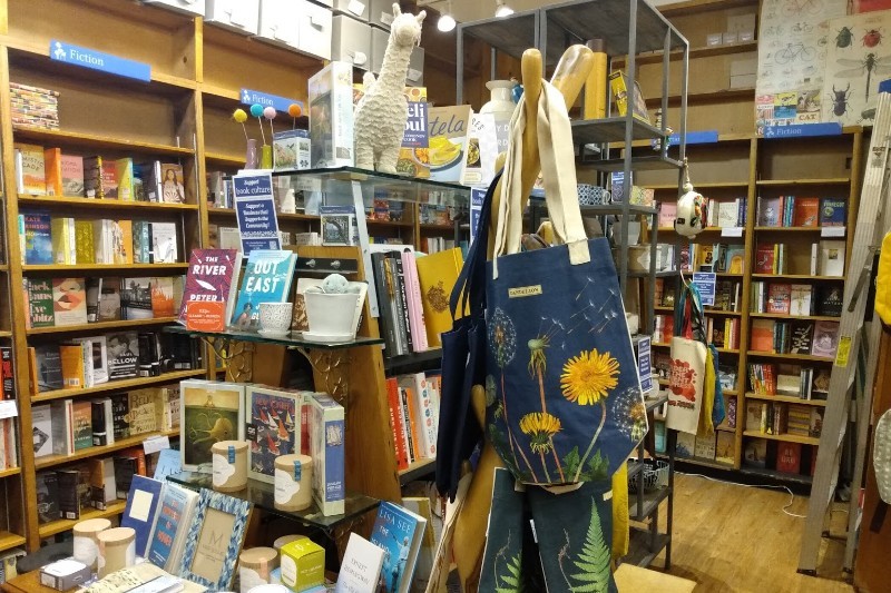A bookstore display with reusable bags hanging to the right of the books. In the background are walls of bookshelves.