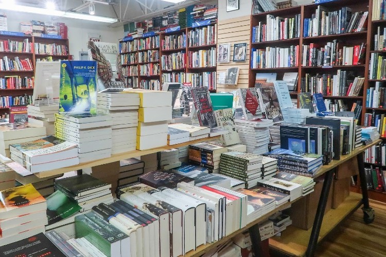 A table piled with books, with wall shelves full of books around it.