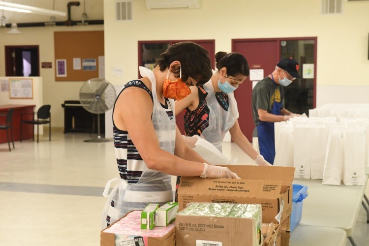 Bagged meal distribution underway at Broadway Community. Photo by Eileen Barroso. A man in a mask and gloves takes food out of a box. In the background are bags for bagged lunches.