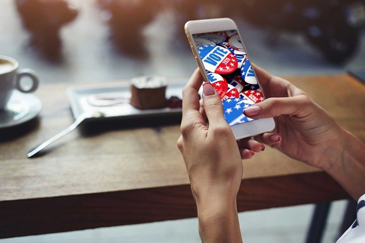 Hands hold a cellphone with image of red, white and blue VOTE buttons; a piece of cake with plate and spoon, plus white cup of expresso lies on a brown table