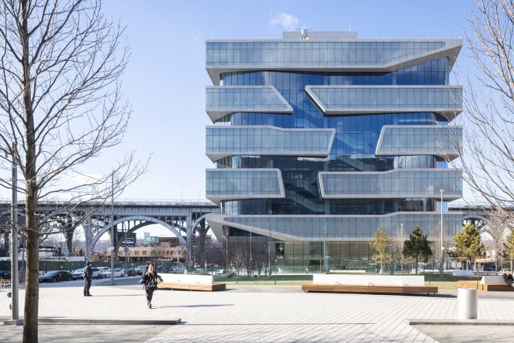 A building against a blue sky with a patio with benches in front. Behind it is the Riverside Drive viaduct.