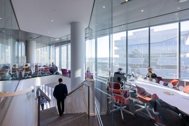 An individual walks down a staircase in a space with large windows showing a contemporary building. There are tables with people sitting at laptop computers.