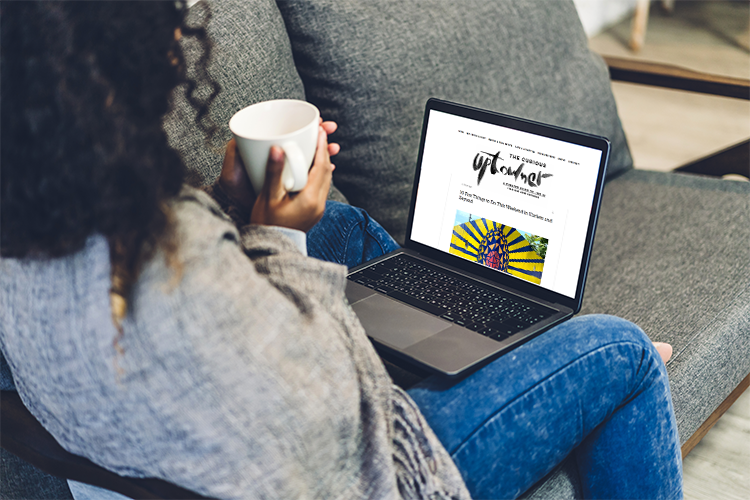 A woman holds a cup of coffee and looks at a laptop with "The Curious Uptowner" on it.