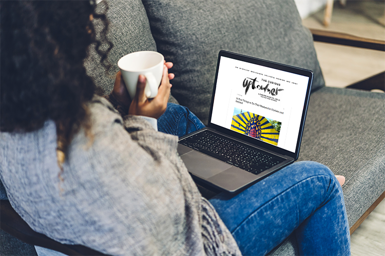 A woman holds a cup of coffee and looks at a laptop with "The Curious Uptowner" on it. 