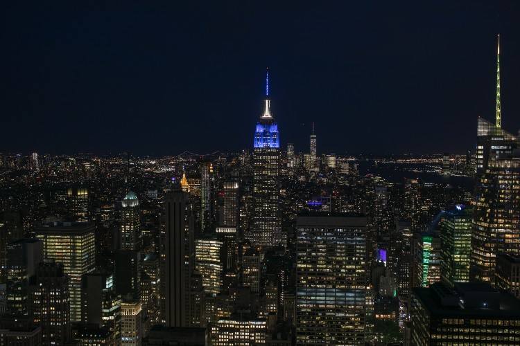The Empire State Building with the New York skyline at night. The building is lit up blue.