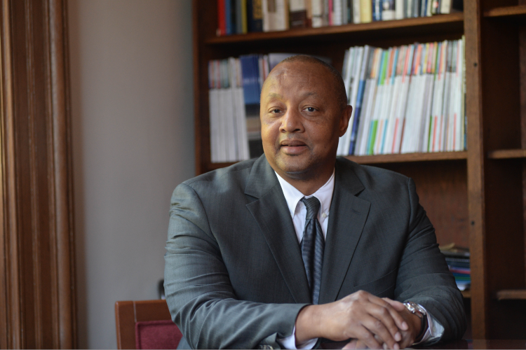 A man in a gray suit sitting in front of a bookcase.