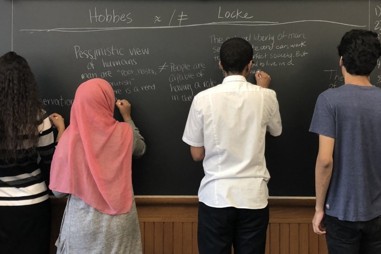 Four students stand at a blackboard writing down the differences between philosophers Hobbes and Locke.