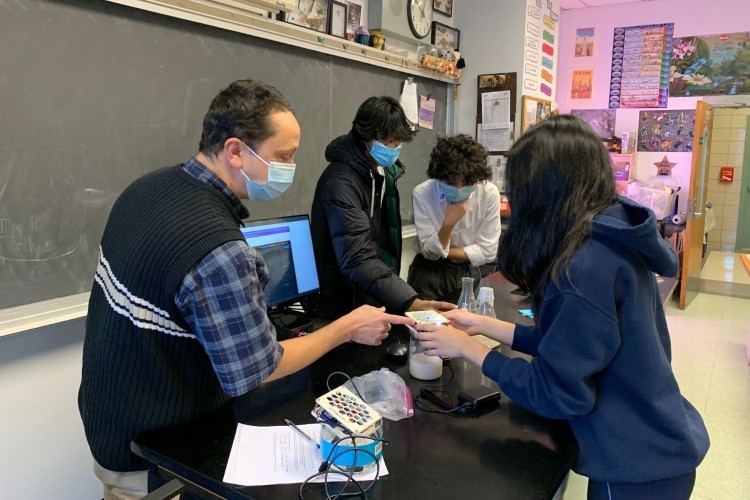 Jason Econome, in his classroom at Stuyvesant High School, overseeing the yeast lab activity he planned this past summer. A group of students standing around a lab table while a teacher shows them an experiment.
