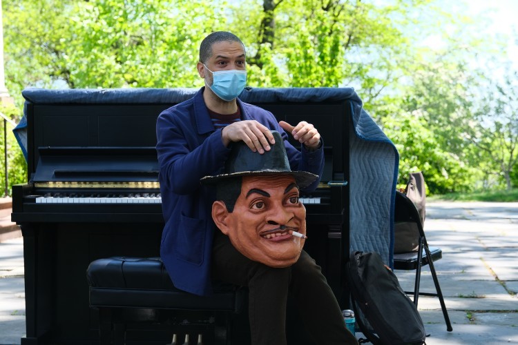 A man sits in front of a piano with a large costume head in his lap.
