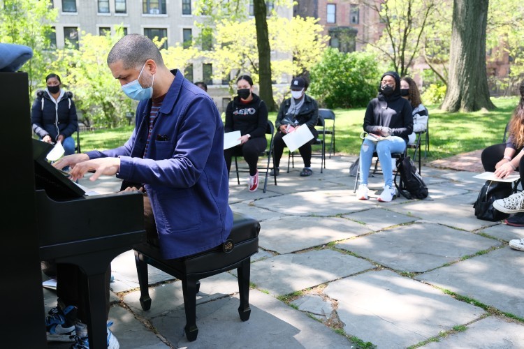 A man plays the piano outdoors in front of student spectators.