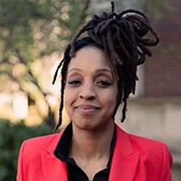 A woman with locs piled on top of her head and wearing a red blazer smiles at the camera.