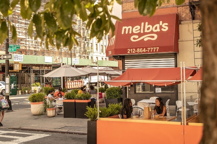 People eating at tables set up on the sidewalk and in the street in front of an awning for Melba's.