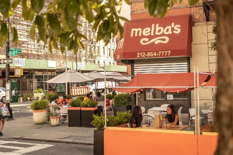 Outdoor dining at Melba's on Frederick Douglass Boulevard. (Photo by Gary He.) People eating at tables set up on the sidewalk and in the street in front of an awning for Melba's.