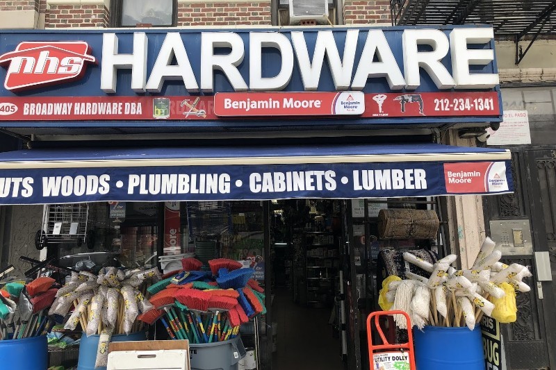 A neon sign saying "nhs Hardware" above a storefront with mops on display on the sidewalk.
