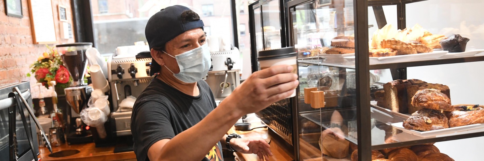 A man in a mask hands a cup of coffee past a display case full of pastries.
