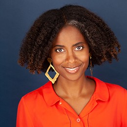A woman with curly hair wearing a bright orange shirt smiles at the camera. 